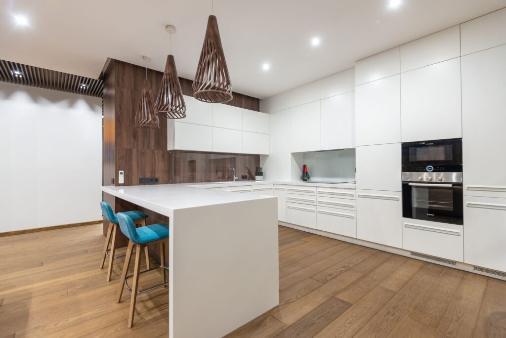 Chairs at table near white cupboards and modern kitchenware with oven in spacious stylish kitchen with hanging lamps in co…