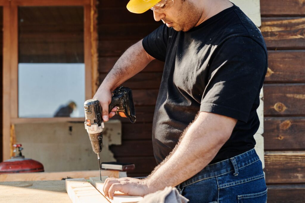 A construction worker wearing a helmet uses a power drill on a wooden plank at an outdoor construction site.