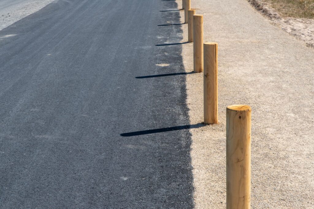 A freshly paved asphalt road bordered by sandy path and wooden posts in bright sunlight.