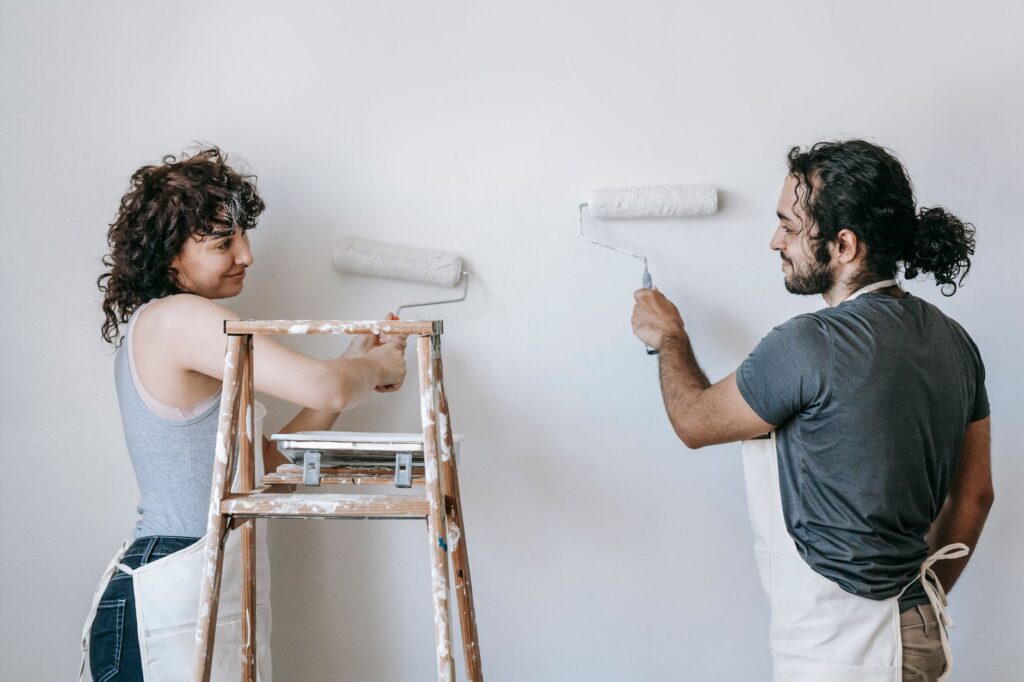 Cheerful adult ethnic couple in aprons looking at each other while painting wall with rollers during renovation process in…