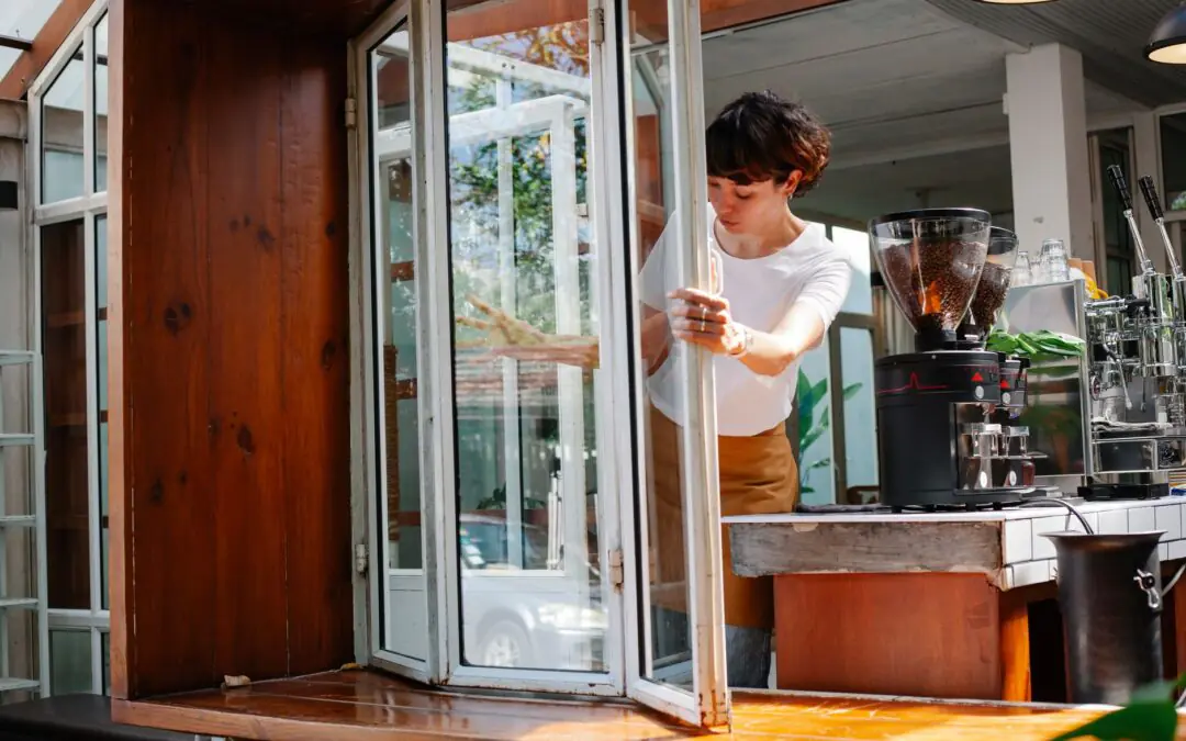 Concentrated female worker in apron opening glass window of modern coffee house with coffee machines during preparation fo…