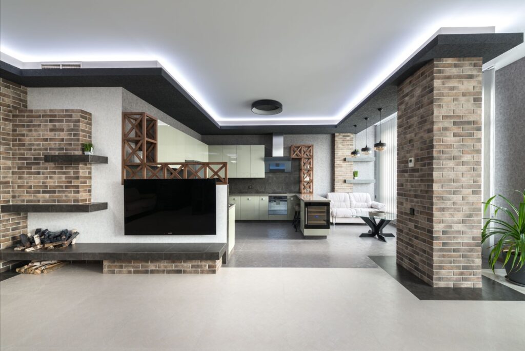 Kitchen with white cupboards placed near couch and modern TV in stylish apartment with pile of firewood and decorative bri…