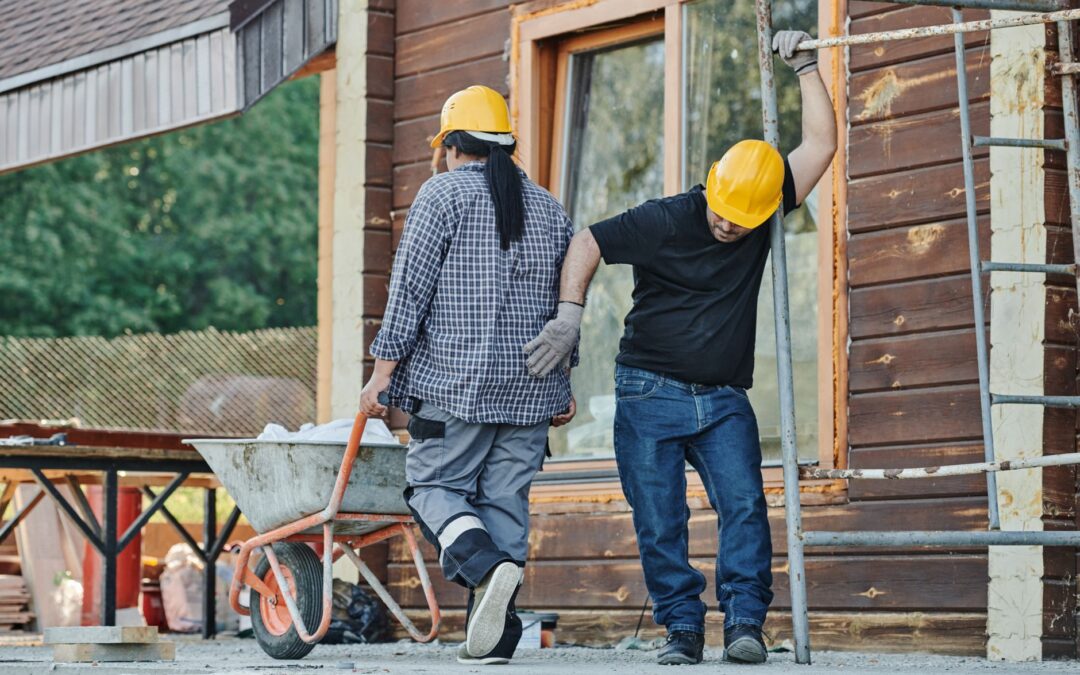 Two construction workers wearing hard hats work at an outdoor site, one with a wheelbarrow.