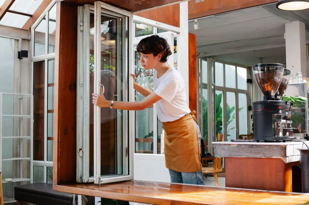 Young waitress in casual clothes and apron standing in modern cafe and looking over window in daytime