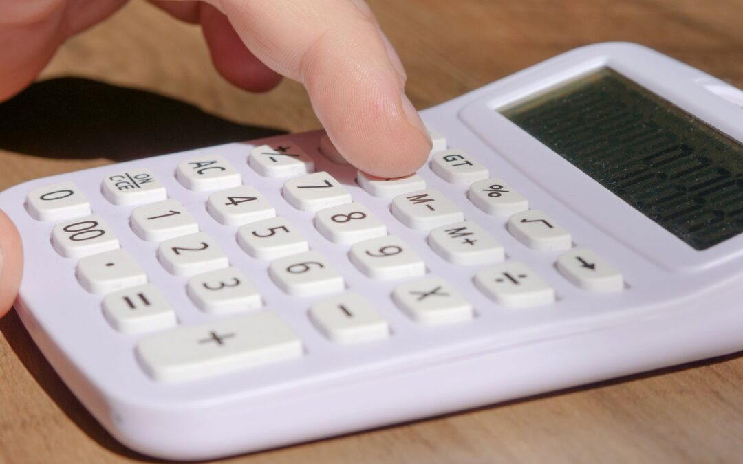A hand pressing keys on a white calculator placed on a wooden desk in bright daylight.