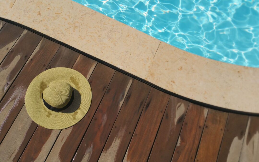 A straw hat rests on a wooden deck beside a sparkling blue swimming pool, perfect for summer relaxation.