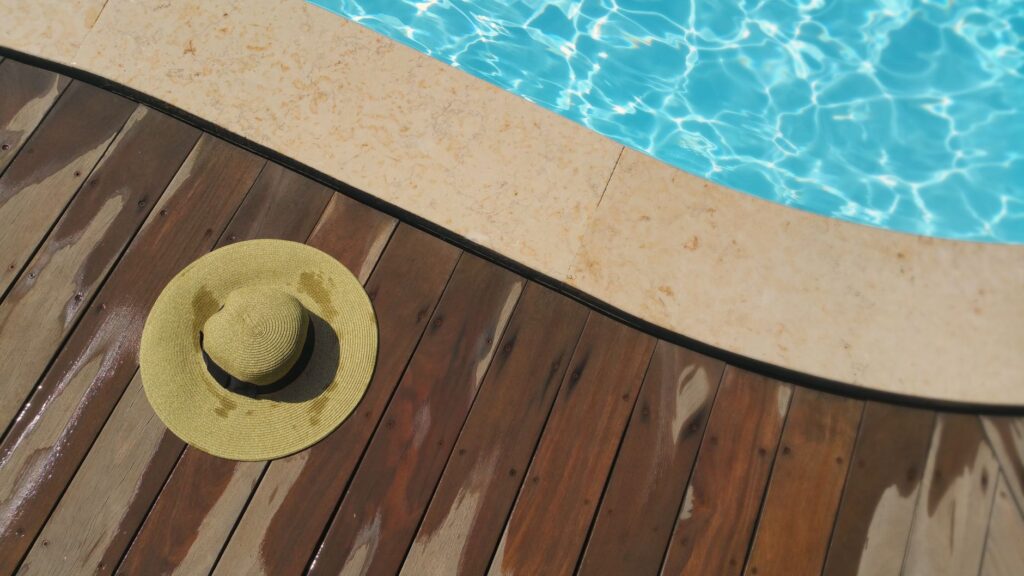 A straw hat rests on a wooden deck beside a sparkling blue swimming pool, perfect for summer relaxation.