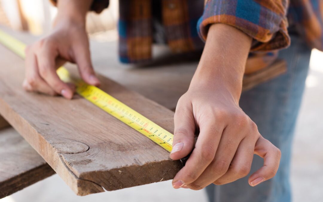 Close-up of hands measuring a wooden plank with a tape measure in a carpentry setting.
