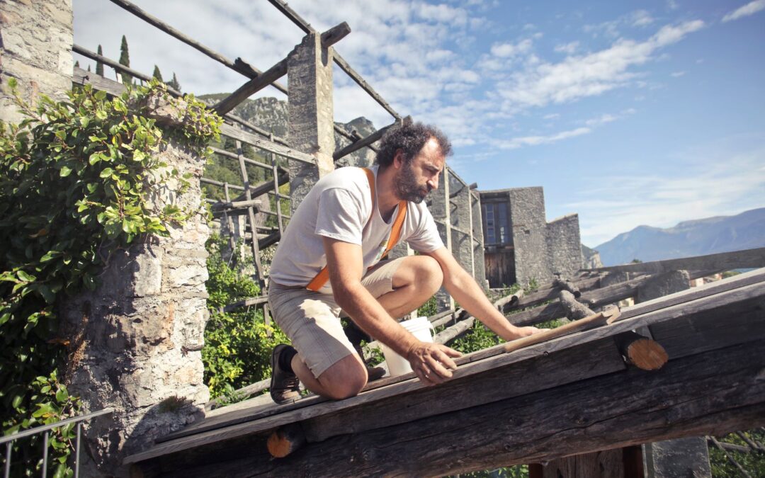 Side view of male builder in summer wear reconstructing rooftop and sitting on wooden beams of aged building on background…
