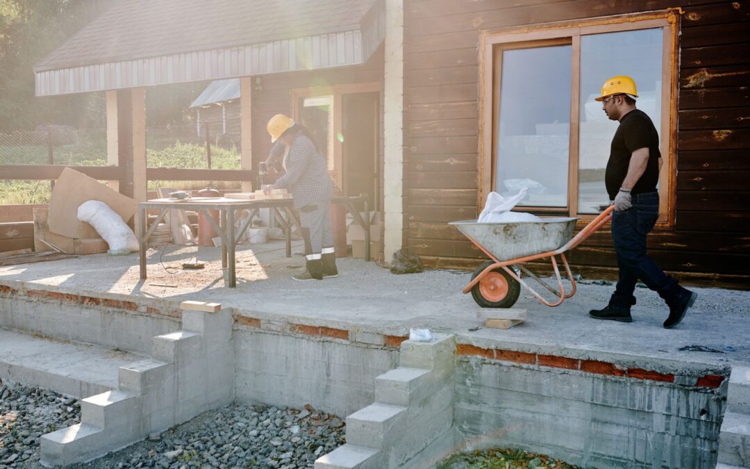 Two construction workers renovating a house exterior, carrying materials and working on woodwork.